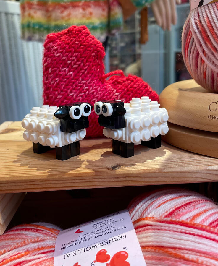 Valentinsschäfchen Zu sehen ist ein Schäfchenpaar aus Klemmbausteinen, der Hintergrund ist dekoriert mit einem gestrickten Herz.